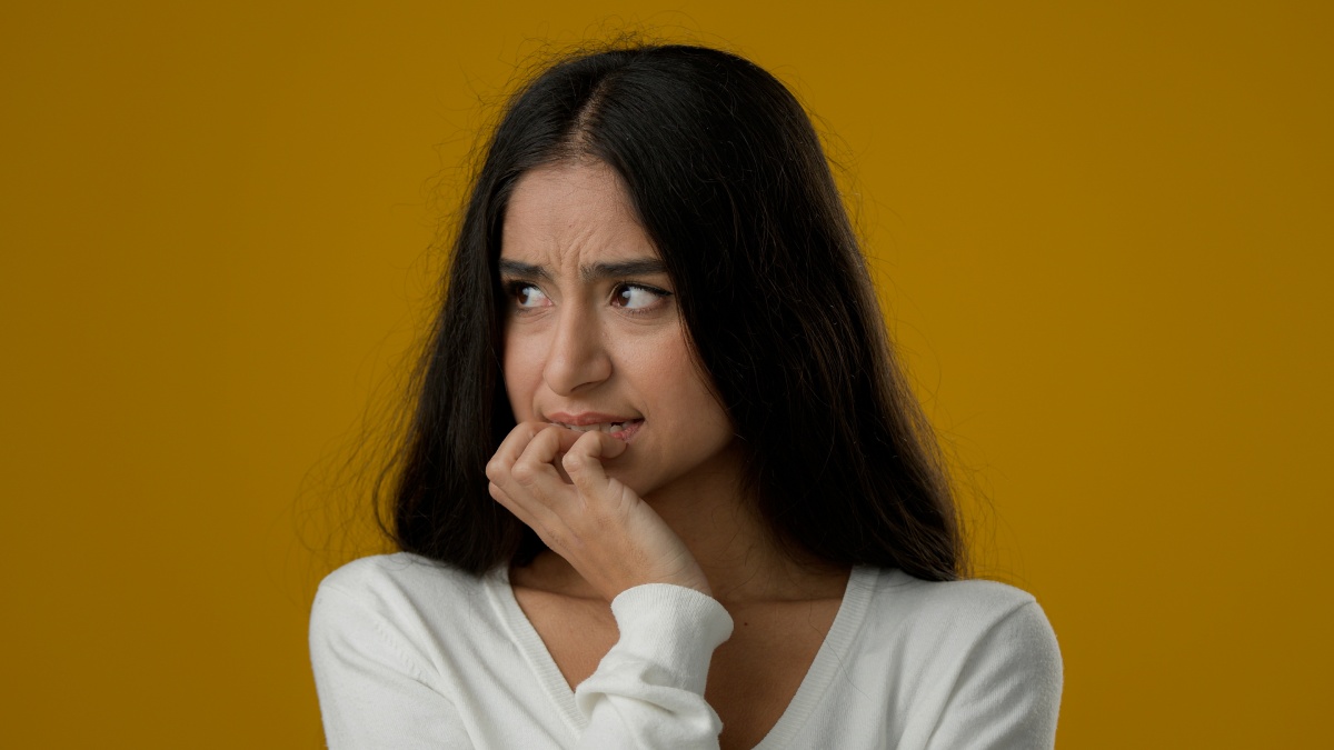 30 something Indian woman nervously chews on fingers while looking to the side against a dark yellow backdrop 